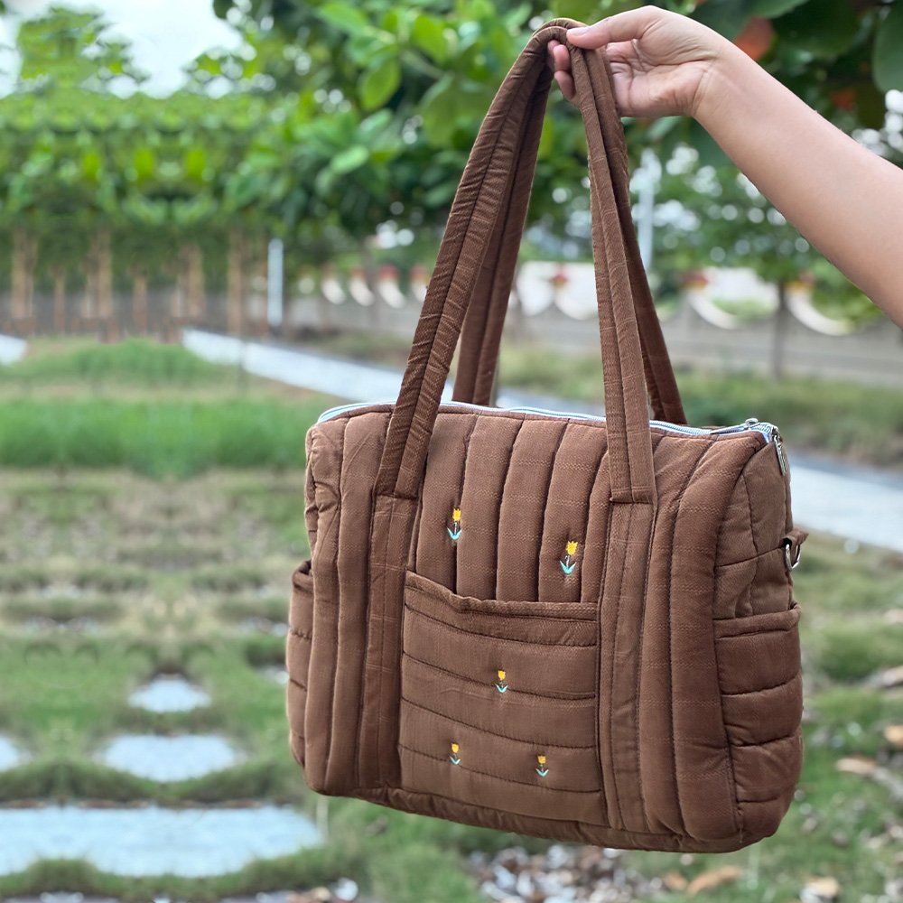 Brown quilted tote bag with front pockets being held by a hand outdoors in a garden setting.