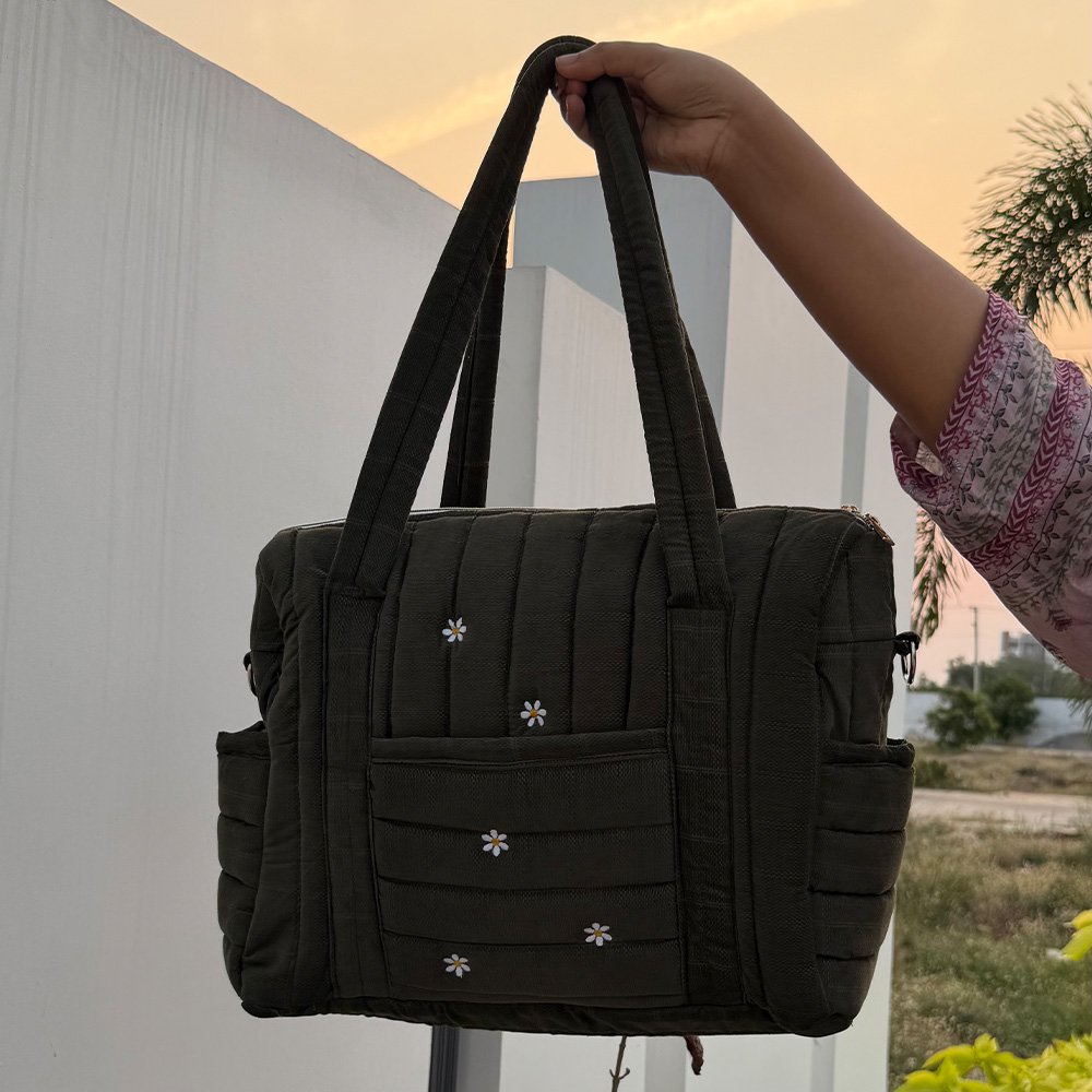 Dark green quilted tote bag with small white daisy embroidery being held by a hand outdoors at sunset