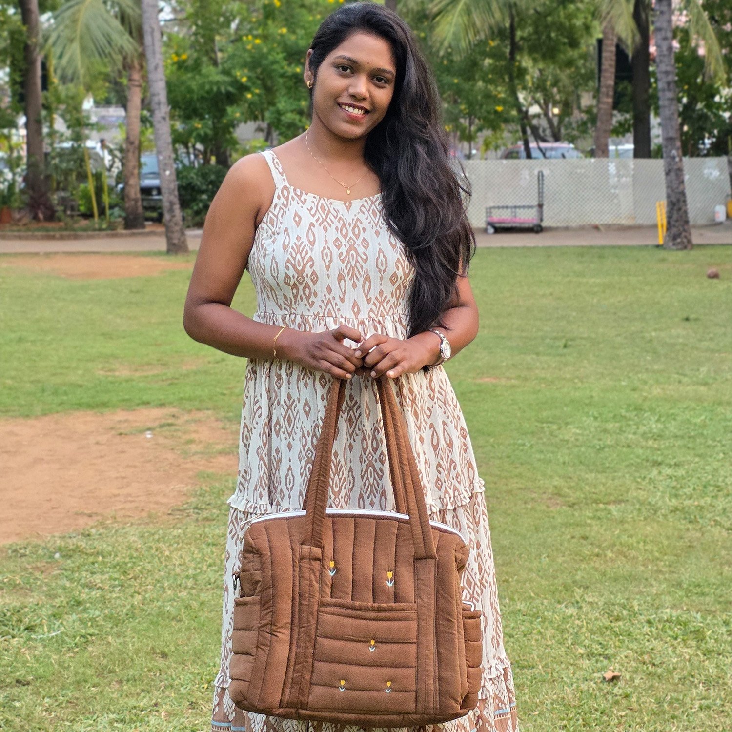 Woman standing in a park, smiling, wearing a cream and brown patterned sundress and holding a brown tote bag in front of her