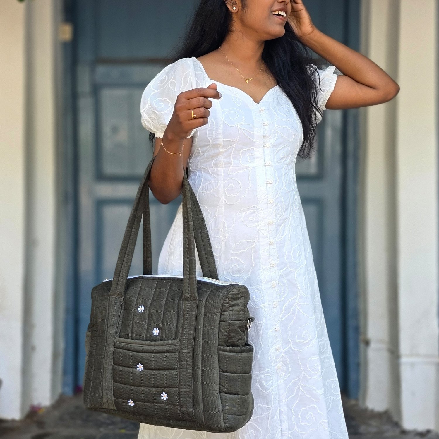 Woman in a white embroidered dress carries a gray quilted tote bag with small white daisies, standing by a doorway.