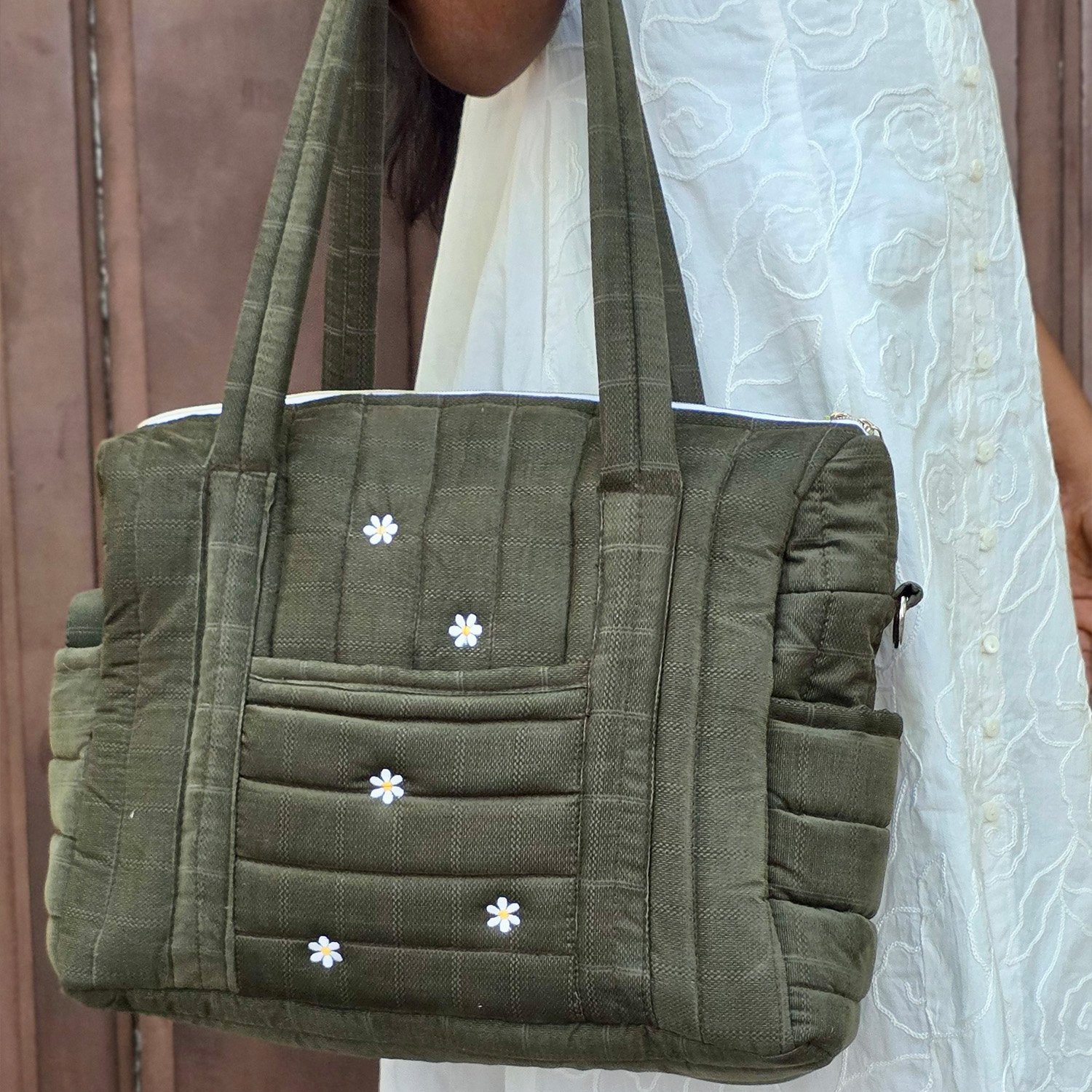 Olive-green quilted handbag with wooden-like fabric and small white daisies embroidered on the front, carried by a person in a white embroidered dress.