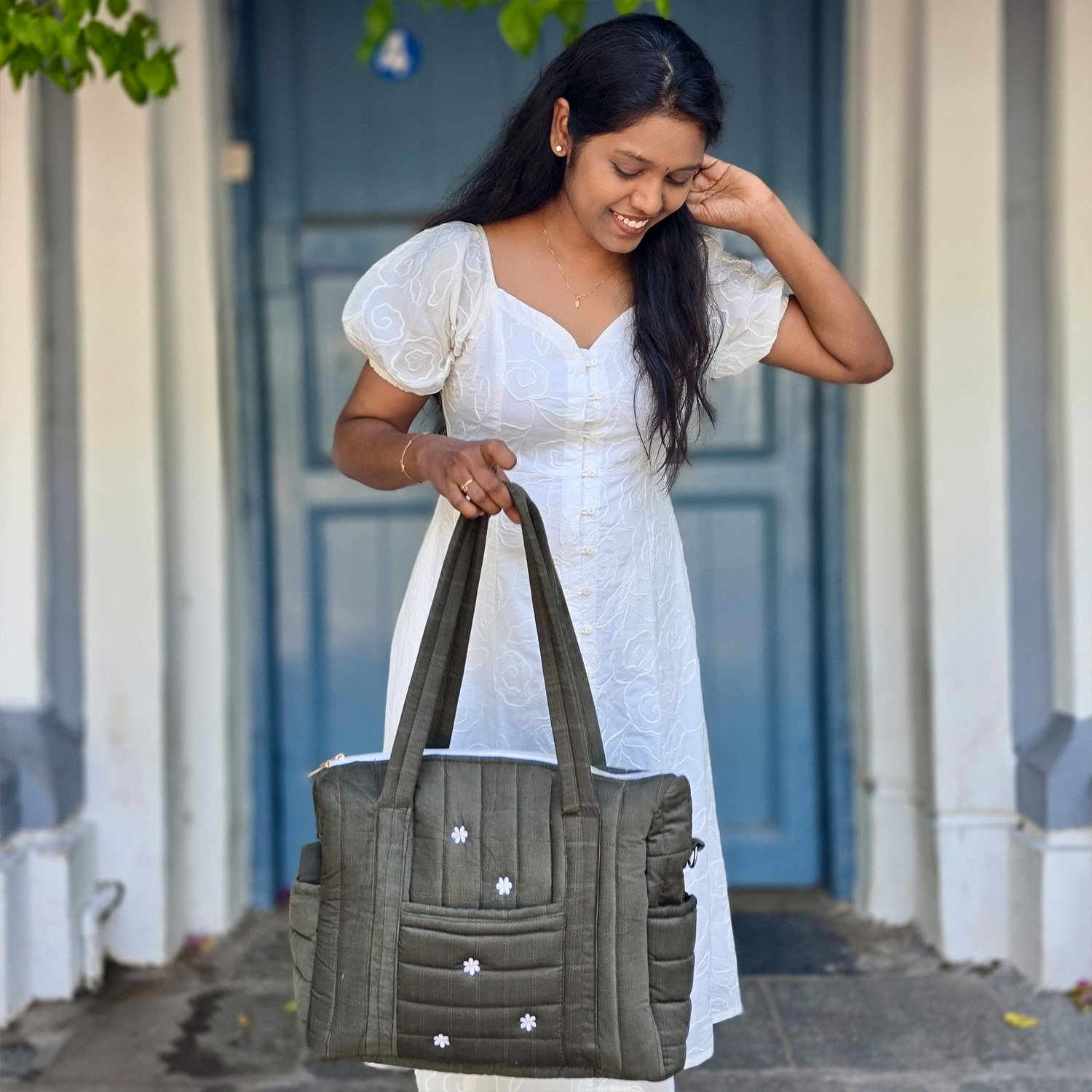 Woman in a white embroidered dress holds a large olive tote bag in front of a blue door. Beyond her, arched entryway and greenery visible.