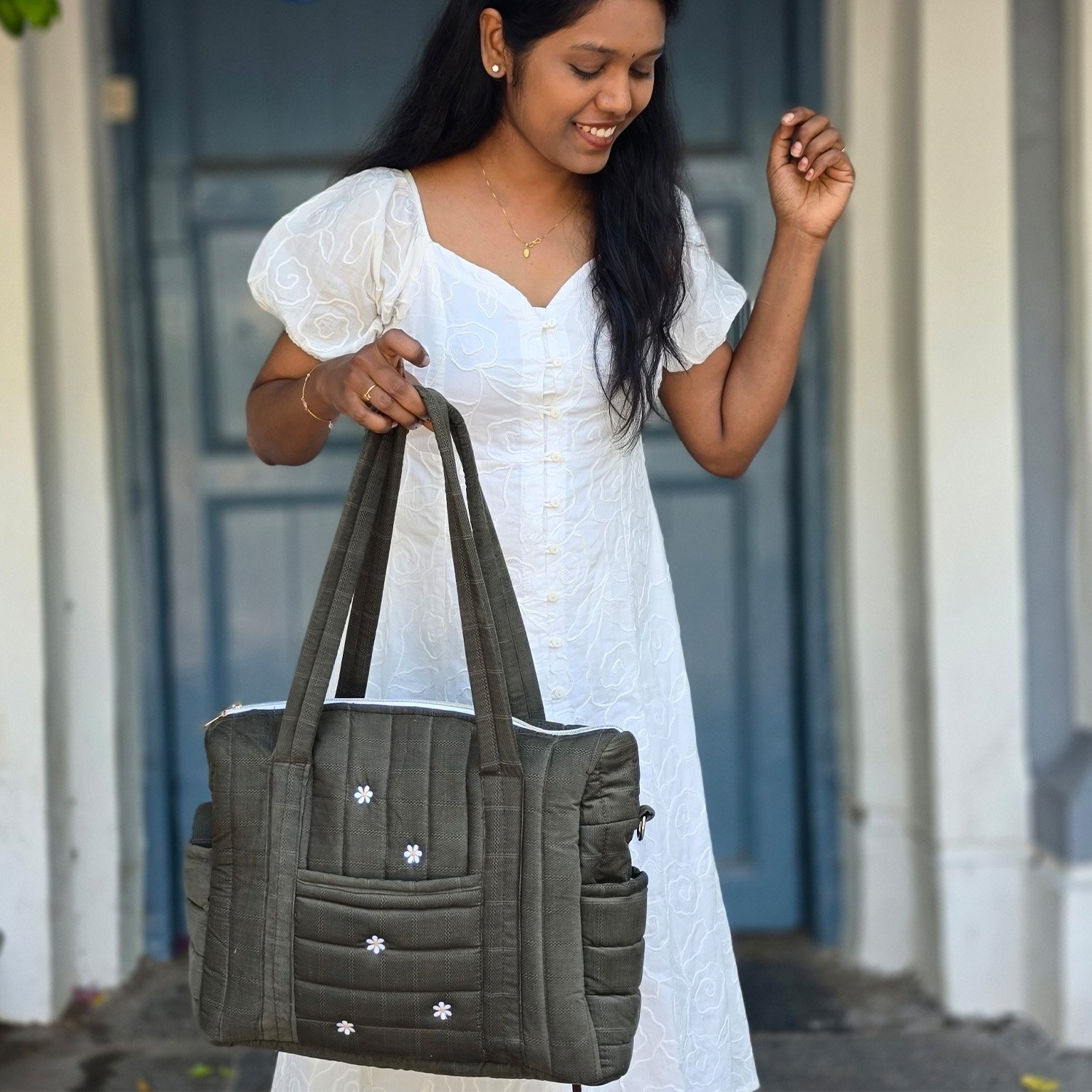 Woman in a white embroidered dress smiles while holding a gray tote bag with small white daisies.
