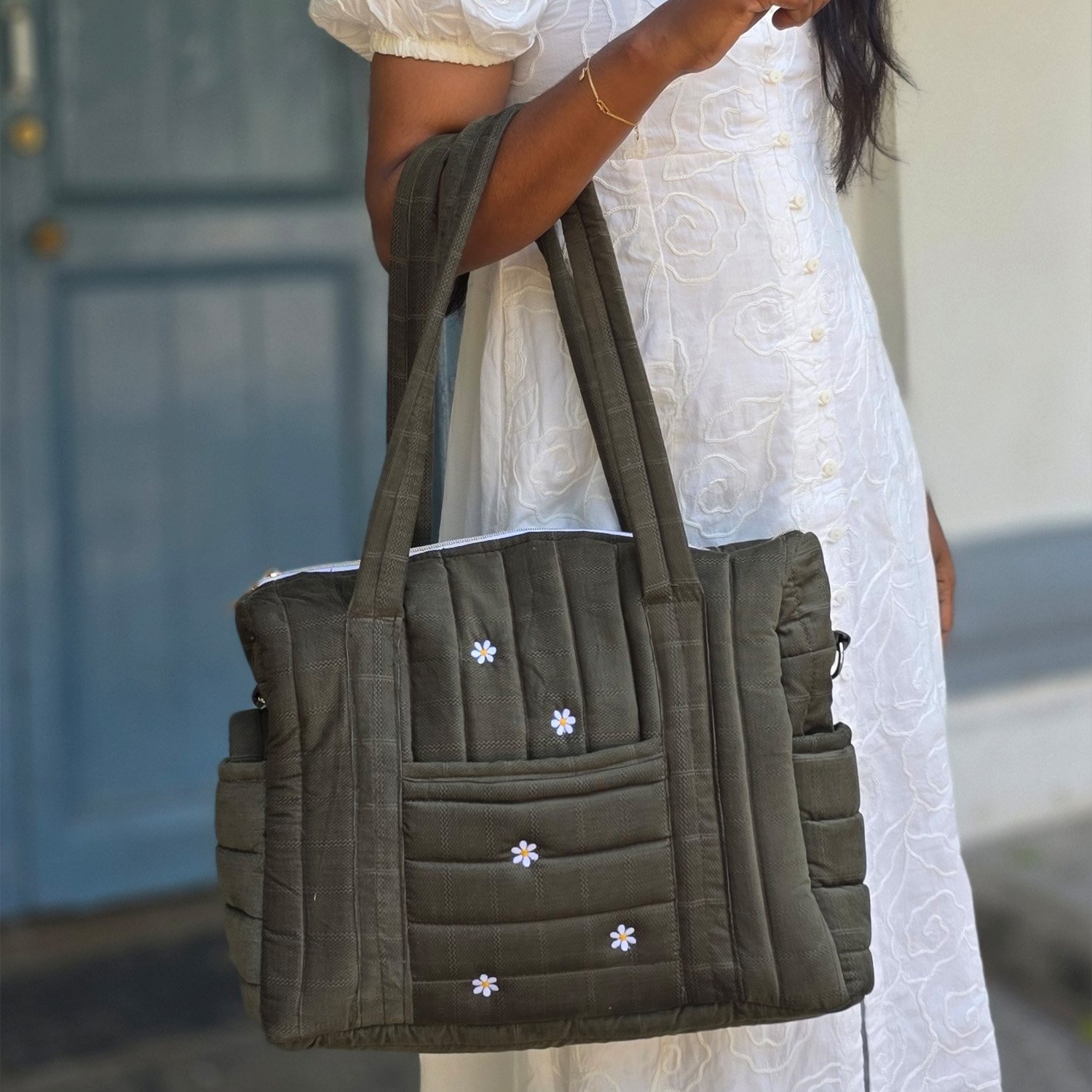 Dark green quilted tote bag with small white daisies carried by a person in a white embroidered dress