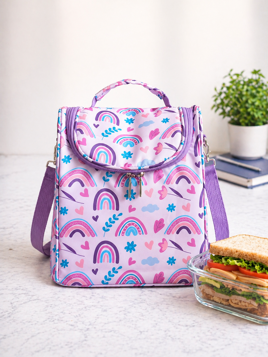 Lavender backpack with pastel rainbow and heart print on a counter, beside a glass lunch container with a sandwich.