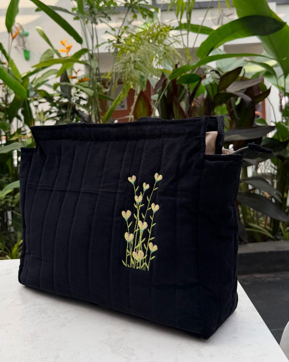 Dark blue quilted tote bag with a small embroidered sprig of heart-shaped flowers in yellow and green, placed on a white surface with green plants in the background