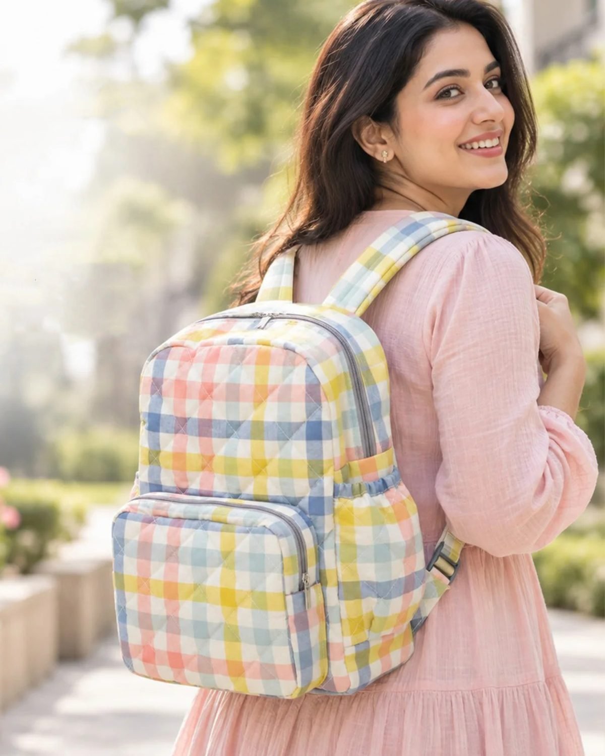 Woman with pastel plaid backpack smiling and looking over her shoulder outdoors.