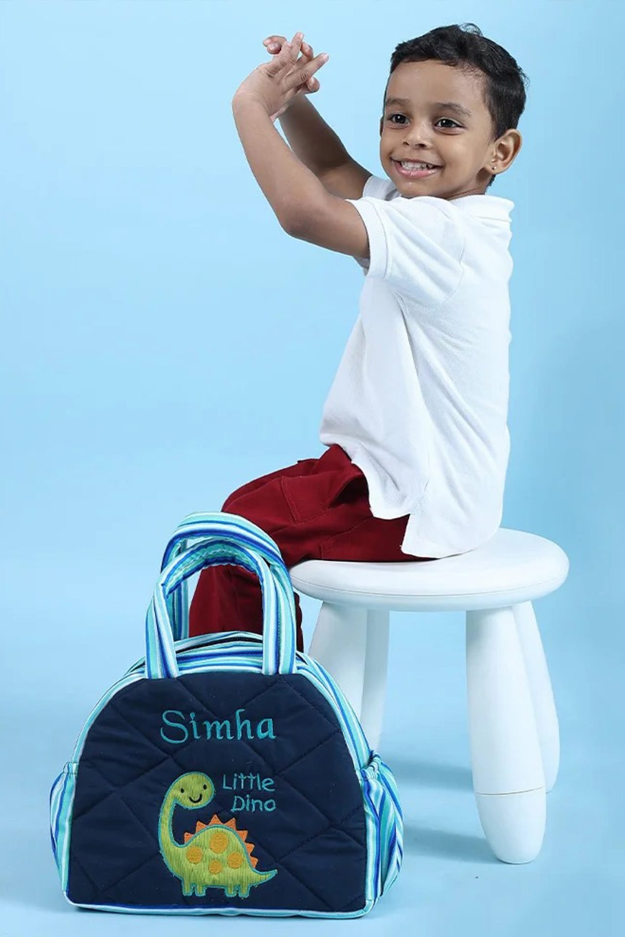 Happy young boy in a white shirt and red shorts sits on a white stool, hands raised, with a blue dinosaur backpack nearby on a light blue background.