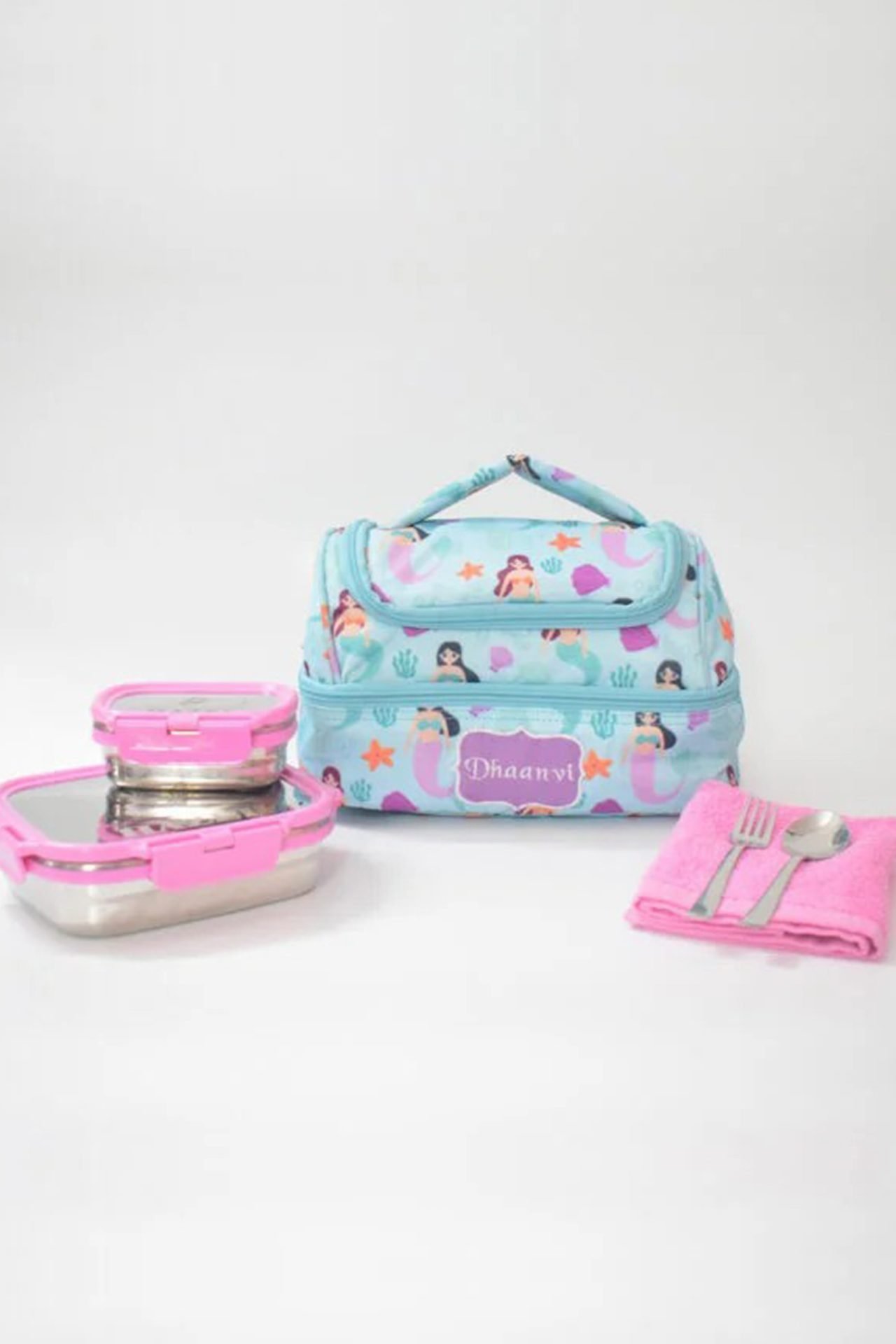 Light blue kids' backpack with mermaid pattern, paired with a pink lunchbox and pink napkin with cutlery on a white background