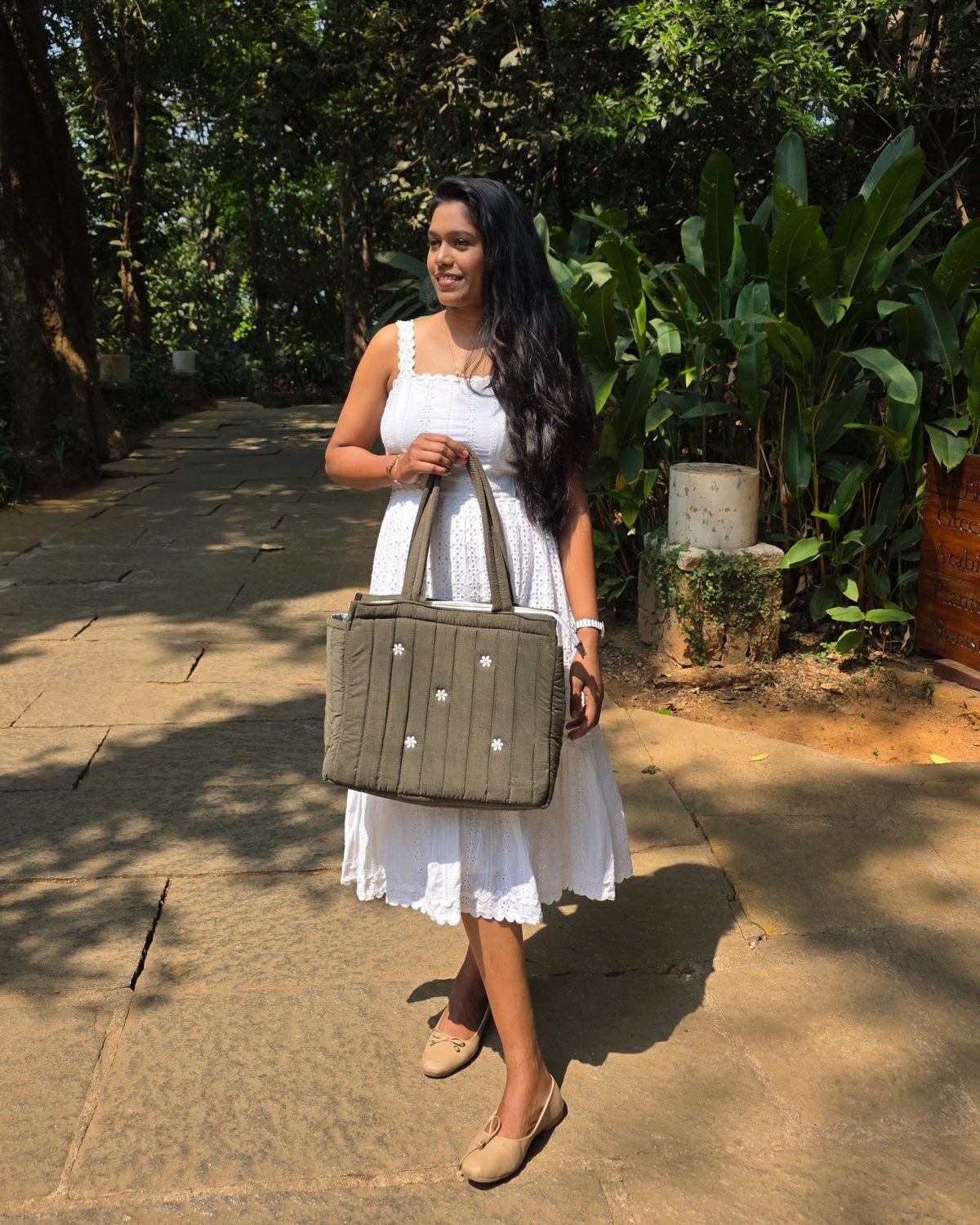 Woman in a white sleeveless dress stands on a stone path, holding a large gray tote bag with small white decorations in a garden setting.
